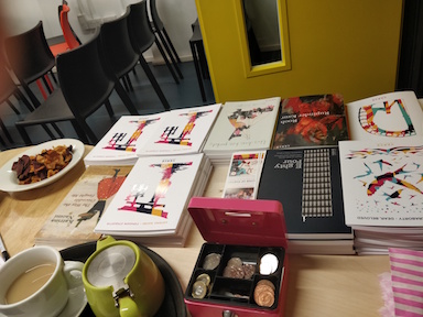 Full colour photo showing what looks like a sales desk covered in piles of books and pamphlets in Verve colours. Also a pink cash box, open, a cup of tea and a green teapot,  some stripey bags, and somebody's dinner on a plate.